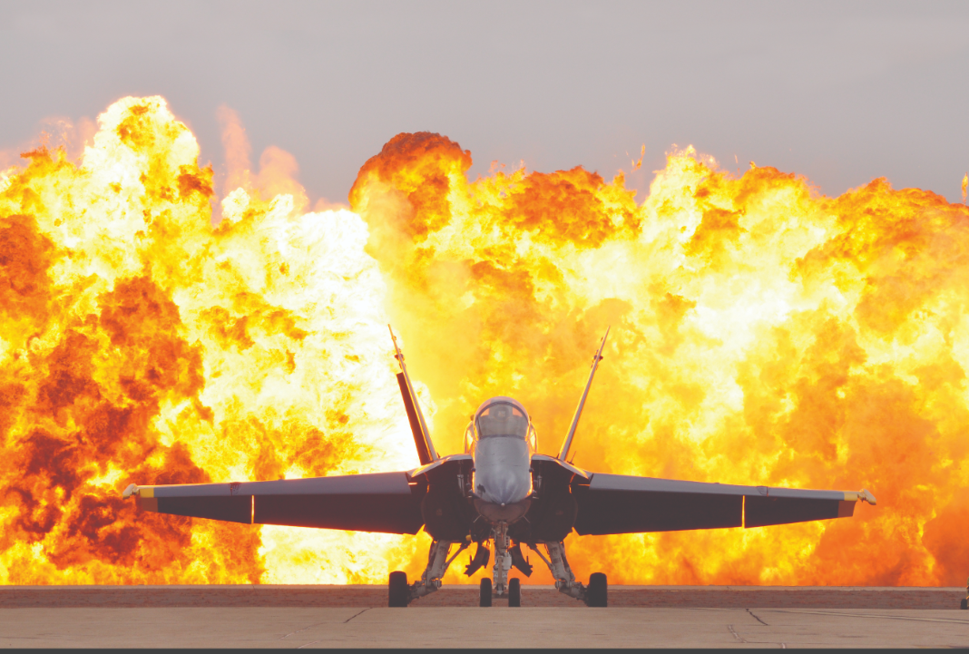 The front view of a jet fighter with explosive flames rising in the background.
